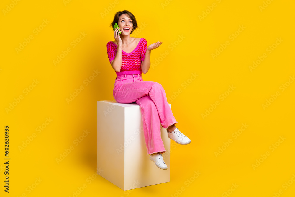 Full length photo of cute woman wear pink knit top sit on cube talk on smartphone look empty space isolated on yellow color background