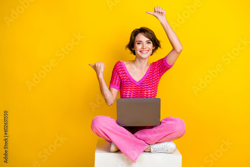 Full size photo of cute girl knit top pink pants indicating empty space sitting on cube with laptop isolated on yellow color background
