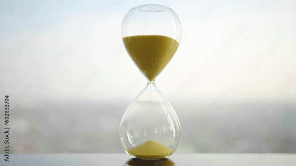 hourglass on table, sand flowing through the bulb of sandglass, time is money concept, times passing concept like work or business deadline, resemble urgency running out of occasion in life