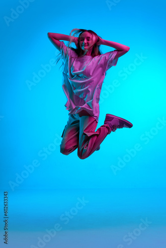 Portrait of pensive, young girl wearing casual clothes jumping in air with closed eyes over blue background in neon light. Levitating dreamer