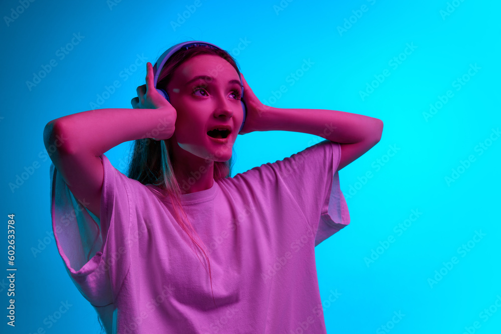 Close up portrait of young girl, teenager in headphones and listening to music with open mouth over blue studio background in neon light Close up portrait of young girl, teenager in headphones and listening to music with open mouth over blue studio background in neon light