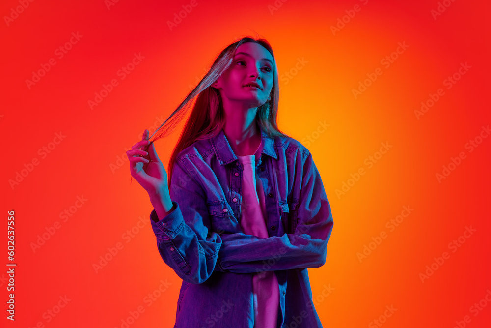 Portrait of young charming girl, teenager with long hair looking away with pensive face over red background in neon light. Concept of beauty, youth