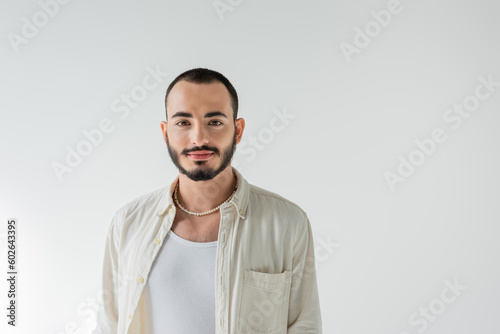 Portrait of young short haired and bearded homosexual man in pearl necklace and casual clothes looking at camera while standing isolated on grey © LIGHTFIELD STUDIOS
