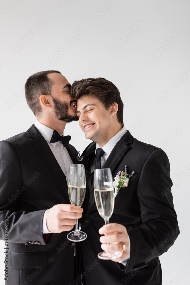 Smiling gay groom in suit with boutonniere whispering to young boyfriend in braces and holding glass of champagne during wedding ceremony isolated on grey Smiling gay groom in suit with boutonniere whispering to young boyfriend in braces and holding glass of champagne during wedding ceremony isolated on grey