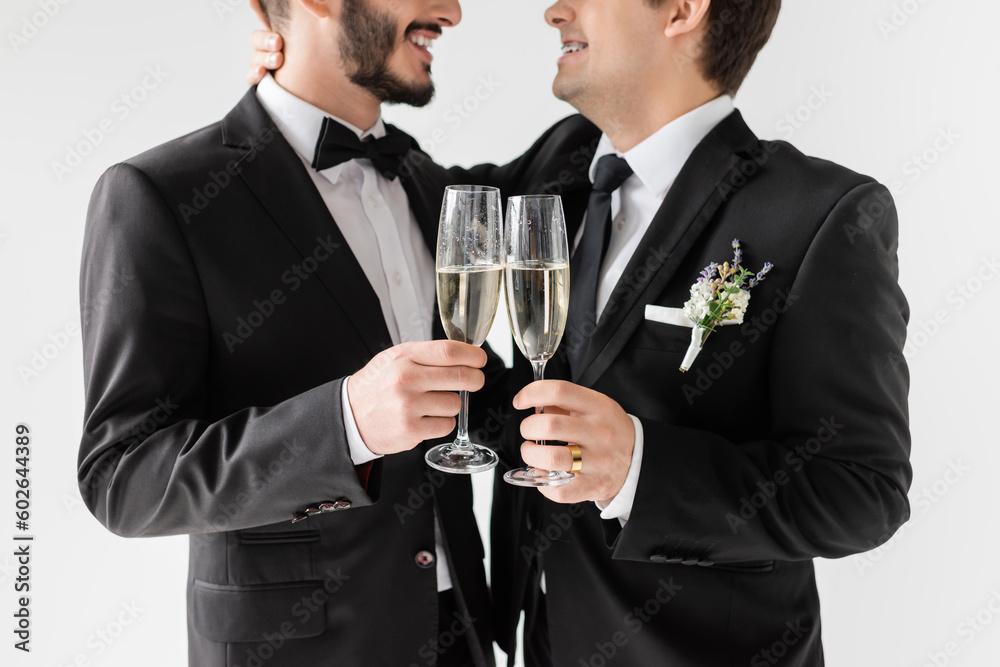 Cropped view of smiling homosexual groom in braces and suit with boutonniere hugging bearded boyfriend and toasting with champagne during wedding ceremony isolated on grey Cropped view of smiling homosexual groom in braces and suit with boutonniere hugging bearded boyfriend and toasting with champagne during wedding ceremony isolated on grey