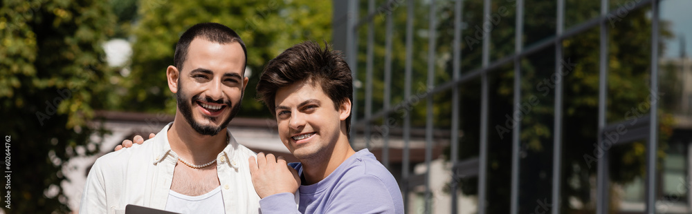 Smiling homosexual man in casual clothes hugging bearded boyfriend and looking at camera together outdoors at daytime, on urban street, banner