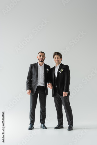 Full length of young and smiling homosexual grooms in classic suits with boutonnieres holding hands and looking at camera on grey background