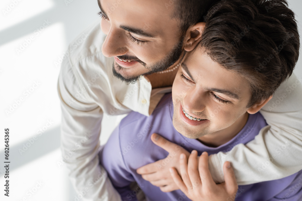 Overhead view of positive and bearded homosexual man hugging young boyfriend in purple sweatshirt and braces on grey background with sunlight Overhead view of positive and bearded homosexual man hugging young boyfriend in purple sweatshirt and braces on grey background with sunlight