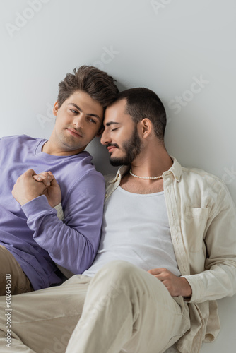 Young homosexual man in sweatshirt holding hand and looking at bearded boyfriend with closed eyes while sitting on grey background