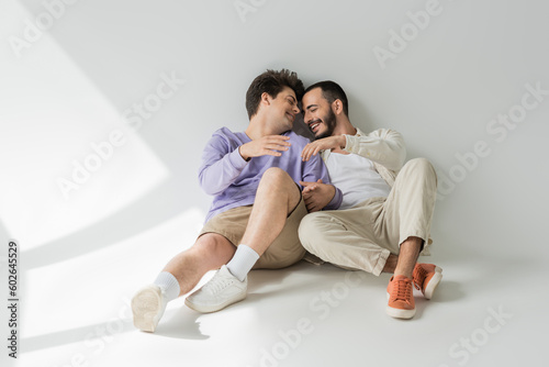 Full length of cheerful same sex partners with closed eyes holding hands while talking and sitting together on grey background with sunlight