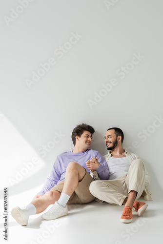 Full length of smiling homosexual couple in casual clothes holding hands and looking at each other while sitting together on grey background with sunlight