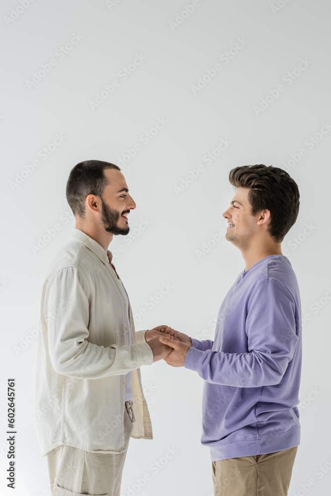 Side view of smiling homosexual couple in casual clothes looking at each other and holding hands while standing isolated on grey Side view of smiling homosexual couple in casual clothes looking at each other and holding hands while standing isolated on grey