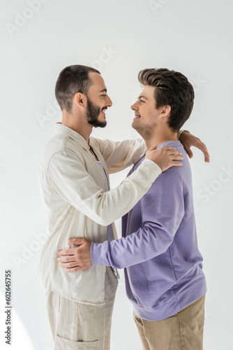 Side view of young and cheerful homosexual couple in casual clothes hugging and looking at each other while standing on grey background