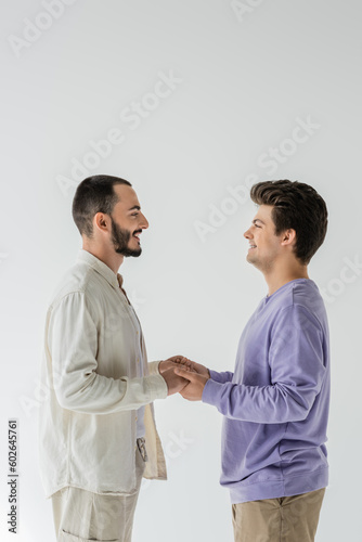 Side view of smiling homosexual couple in casual clothes looking at each other and holding hands while standing isolated on grey
