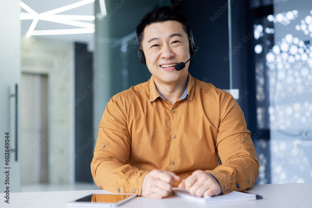 Cheerful and smiling online customer support tech worker, Asian man with call headset smiling and looking at webcam, working on helpline and consulting customers, using headset
