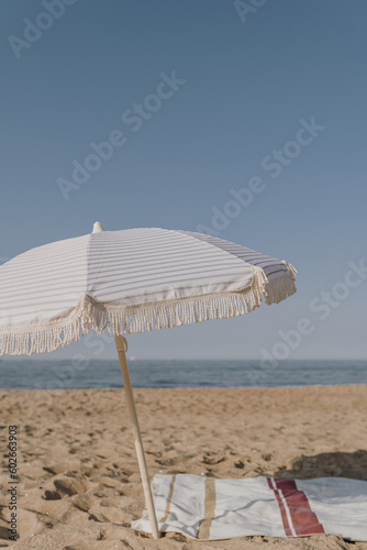 Minimal summer holidays vacation concept. Beach umbrella, beach towel on sand in front of blue sky and sea. Chilling, lounging on the beach