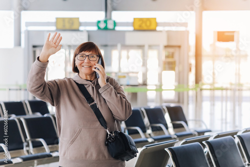 Happy smiling mature traveler woman hold smartphone and wave hand in greeting someone who meeting her at airport terminal after arrived from trip at new country at holiday vacation. Retirement travel