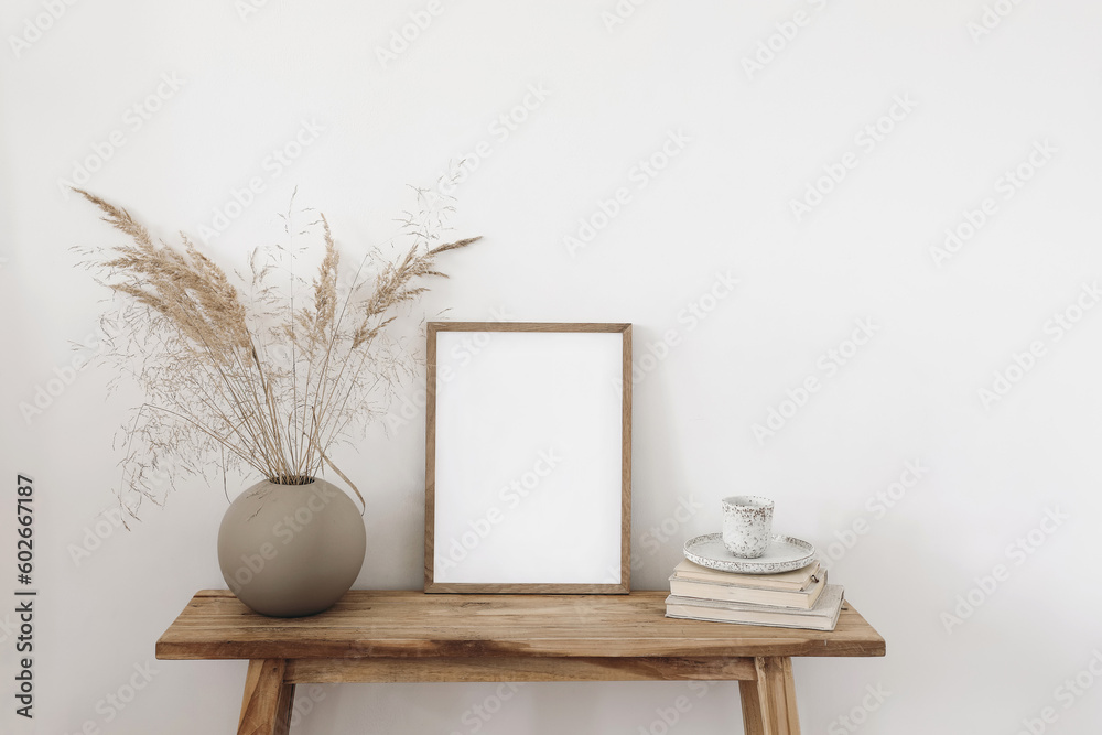 Ball shaped vase with dry grass bouquet. Old wooden bench. Blank vertical picture frame mockup with books and cup of coffee, tea. White wall background. Empty copy space. Neutral still life interior. - obrazy, fototapety, plakaty 