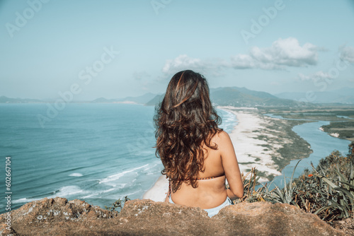 Mulher posando para foto no alto do morro, com paisagem da praia ao fundo. Espaço para texto.