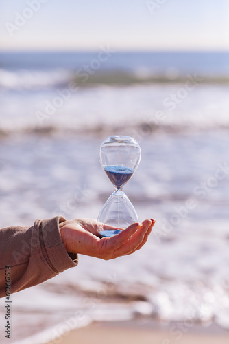 Time is running fast. Hourglass with blue sand inside in mature woman's hand symbolize the brevity of life. Background is sea with beautiful sunlights. Concept of the rapid passage of time. Copy space