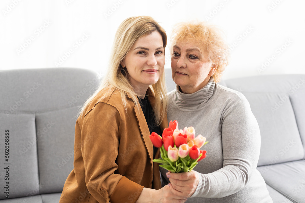 Beautiful senior mom and her adult daughter are hugging and smiling while sitting on couch at home Beautiful senior mom and her adult daughter are hugging and smiling while sitting on couch at home
