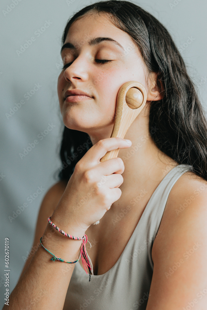 Young woman applying facial massage with wooden roll. Applying pressure to improve circulation. New methods of skincare and daily rituals. Relaxed selfcare day.Portrait in studio with grey background. Young woman applying facial massage with wooden roll. Applying pressure to improve circulation. New methods of skincare and daily rituals. Relaxed selfcare day.Portrait in studio with grey background.