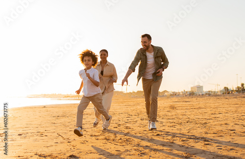 Beach fun. Happy diverse family walking and fooling by the seaside, boy running along beach being chased by his parents