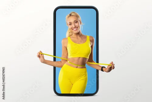 Obraz Positive young european slim woman in sportswear measures waist with measuring tape on smartphone