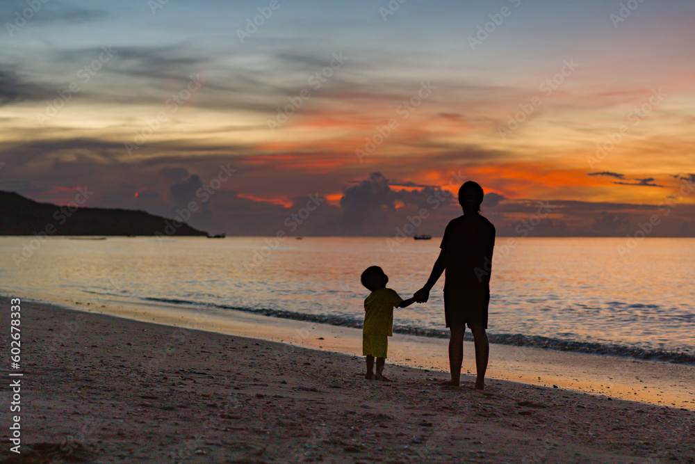 Mother and her little child walking down the beach park holding hands at sunset