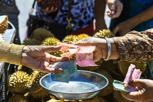 Elderly Thai female vendor ride motorcycle selling durians and fruits and vegetables to tourists at roadside. Street Merchant cut open and sell flesh of fresh durian. Durian trading. Selective focus.