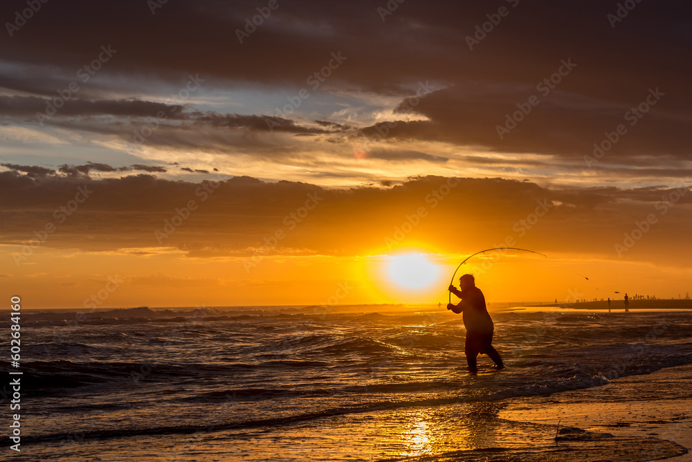 Silhouette of a man fishing on the seashore at the beach during sunset.