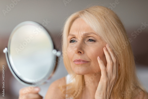 Upset senior woman checking wrinkles around her eyes, looking in mirror and touching face, making beauty routine at home