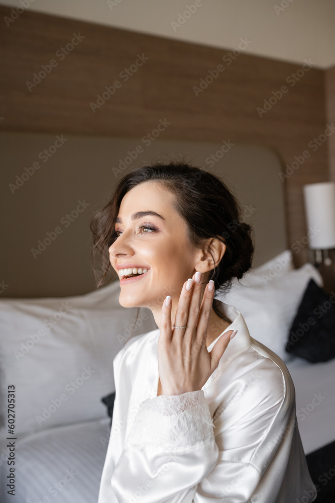 excited young woman with brunette hair in white silk robe showing engagement ring on finger and smiling in hotel room on wedding day, special occasion, young bride excited young woman with brunette hair in white silk robe showing engagement ring on finger and smiling in hotel room on wedding day, special occasion, young bride