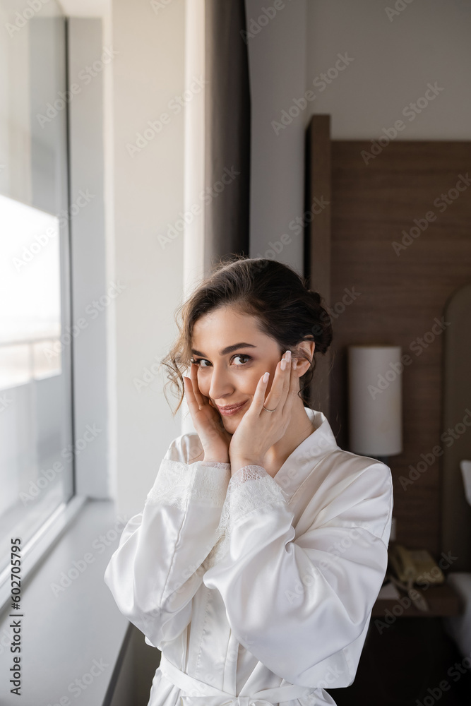 cheerful young bride with engagement ring on finger touching cheeks and standing in white silk robe next to window in hotel suite, special occasion, bride on wedding day cheerful young bride with engagement ring on finger touching cheeks and standing in white silk robe next to window in hotel suite, special occasion, bride on wedding day