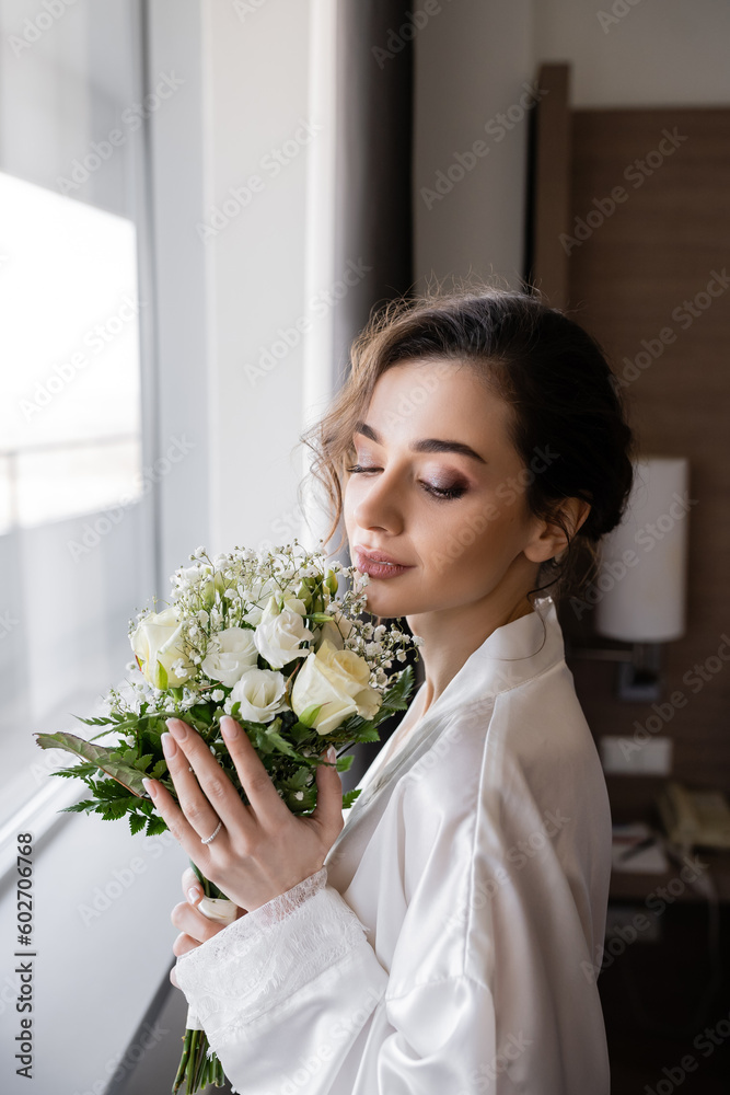 young woman with engagement ring on finger standing in white silk robe and looking at floral bridal bouquet next to window in hotel suite, special occasion, bride on wedding day young woman with engagement ring on finger standing in white silk robe and looking at floral bridal bouquet next to window in hotel suite, special occasion, bride on wedding day