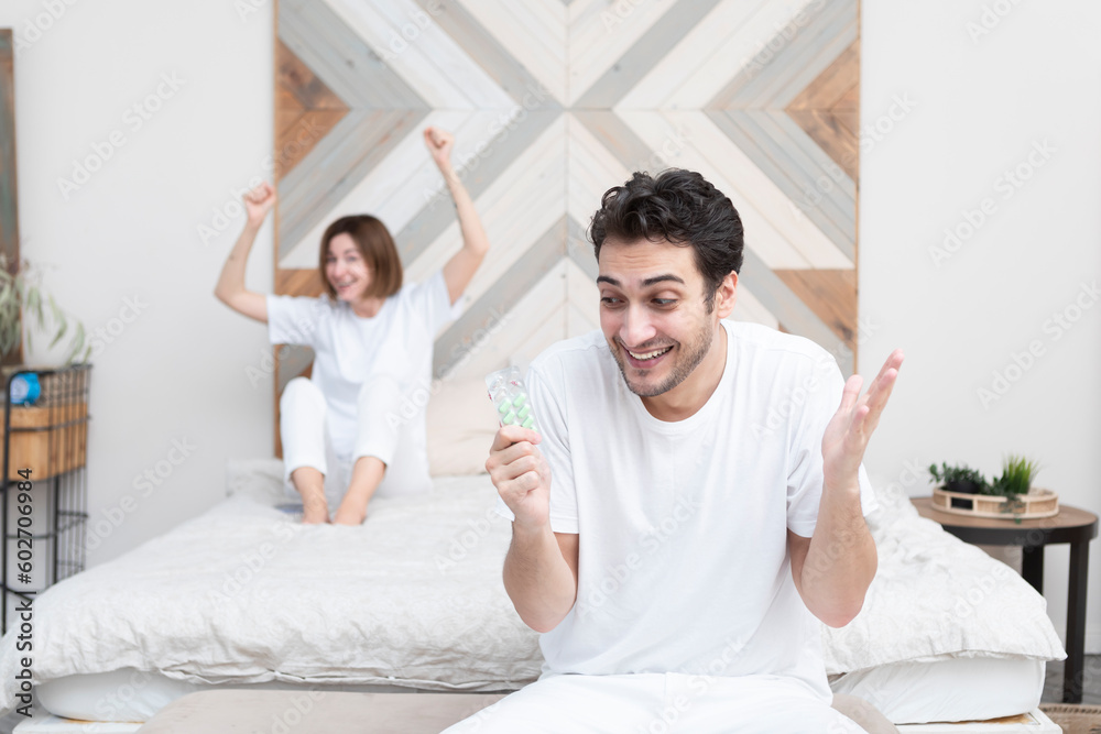 Obraz premium Happy man sitting on the bed, holding tablets and demonstrating his happiness after solving his erectile dysfunction problem