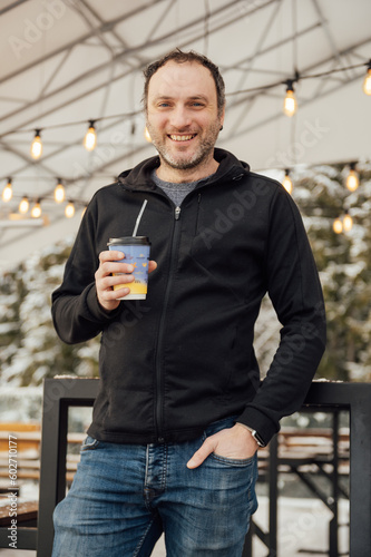 A handsome guy in a black sweater stands with a glass of coffee against the backdrop of mountains and edison lamps