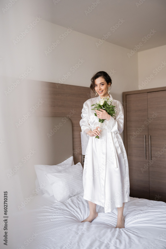 cheerful young bride with brunette hair in white silk robe and pearl earrings holding bridal bouquet while standing on bed in hotel room, special occasion, happy woman