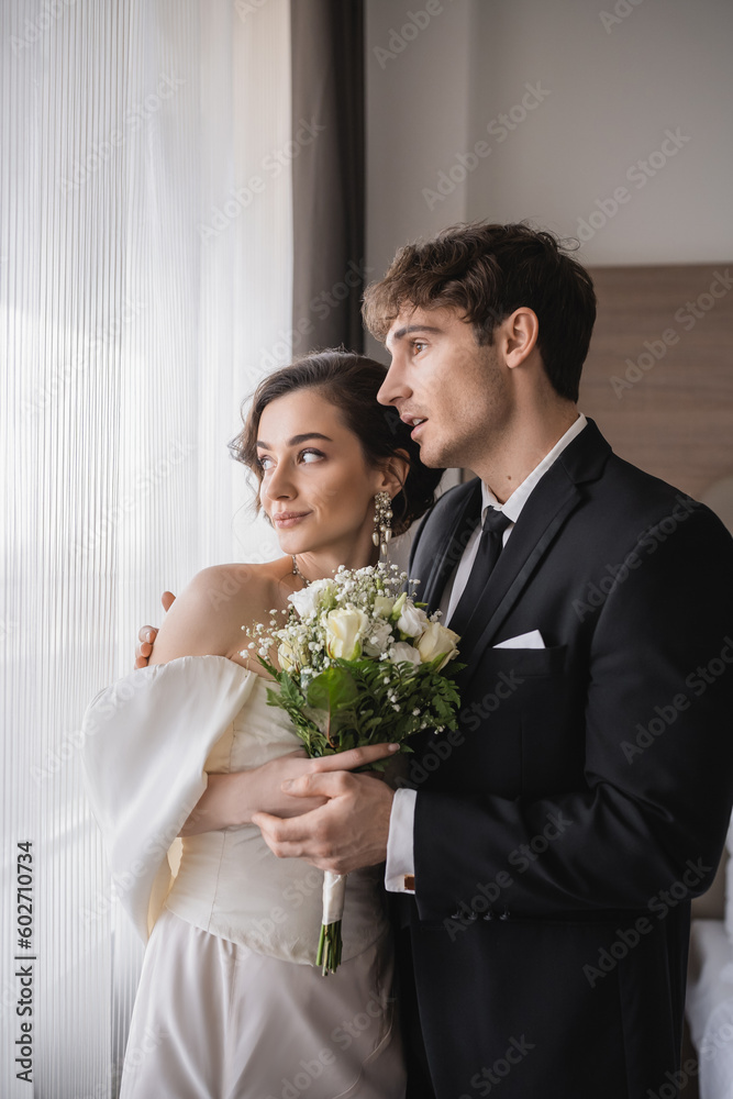 amazed groom in classic formal wear standing with opened mouth next to elegant young bride in jewelry, white dress with bridal bouquet in modern hotel room after wedding ceremony amazed groom in classic formal wear standing with opened mouth next to elegant young bride in jewelry, white dress with bridal bouquet in modern hotel room after wedding ceremony