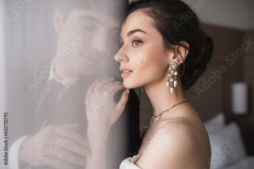 stunning bride in elegant jewelry and wedding dress hugging shoulder of groom in classic formal wear while standing together behind white tulle in modern hotel room after ceremony