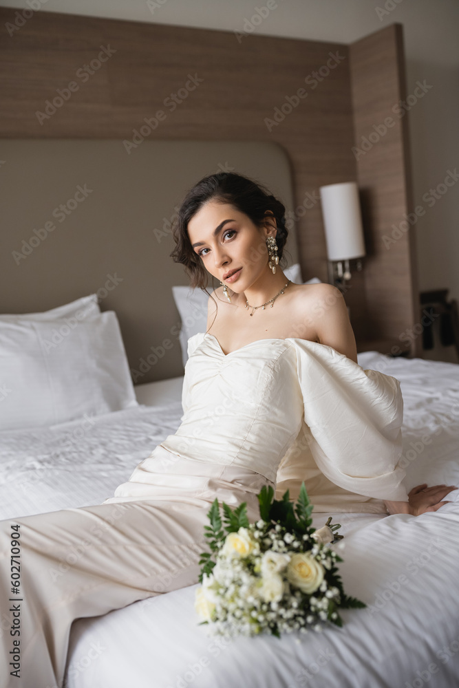 stunning young bride in white dress and luxurious jewelry sitting on bed next to bridal bouquet with flowers and looking at camera in modern bedroom in hotel room on wedding day stunning young bride in white dress and luxurious jewelry sitting on bed next to bridal bouquet with flowers and looking at camera in modern bedroom in hotel room on wedding day