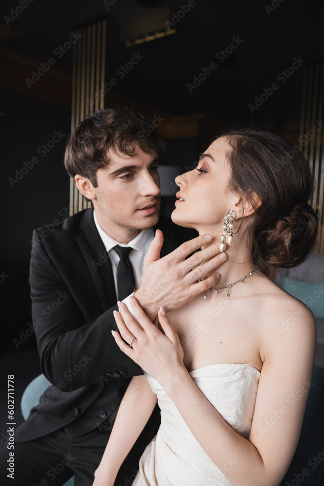 groom in black suit with tie touching face of charming bride in white wedding dress and jewelry while looking at each other in hotel room, couple on wedding day