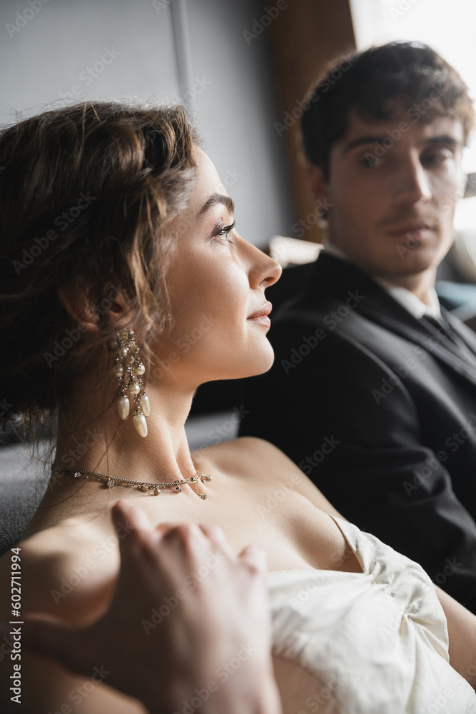 elegant bride in white wedding dress and luxurious jewelry looking away near handsome groom in suit looking at her on blurred background of hotel room elegant bride in white wedding dress and luxurious jewelry looking away near handsome groom in suit looking at her on blurred background of hotel room