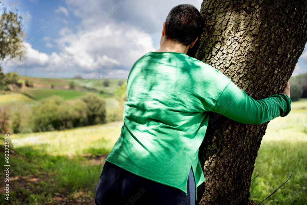 Non-binary woman with very short hair hugging a large oak tree in a field, green T-shirt and hair that is beginning to graying. Non-binary woman with very short hair hugging a large oak tree in a field, green T-shirt and hair that is beginning to graying.