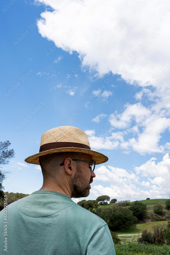 portrait of a man in his forties with glasses and beard, wearing an unrecognizable straw hat, looking at the horizon with a sky with white clouds and a green and wet field.