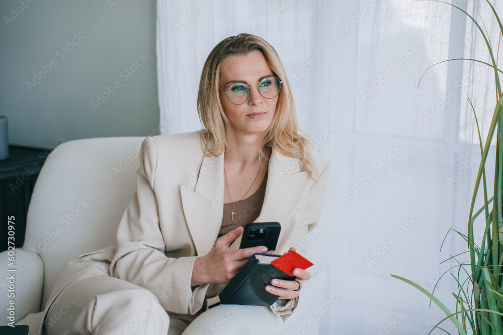 Pensive blonde caucasian businesswoman in beige suit sitting on cozy chair looks away holds phone, credit card and diary, pulled by financial crisis, troubles. Young women trying pay bills. Pensive blonde caucasian businesswoman in beige suit sitting on cozy chair looks away holds phone, credit card and diary, pulled by financial crisis, troubles. Young women trying pay bills.