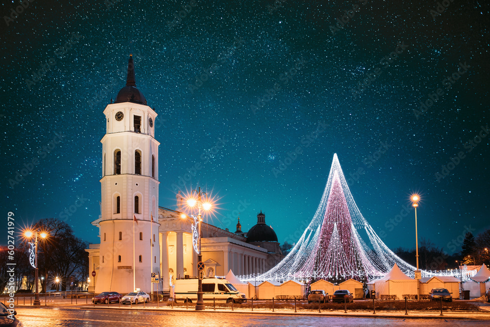 Obraz Vilnius, Lithuania. Amazing Bold Bright Blue Starry Sky Gradient Above Christmas Tree On Background Bell Tower Belfry Of Vilnius Cathedral At Square In Evening New Year Christmas Xmas Illuminations. fototapeta, plakat