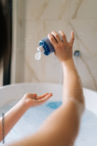 Vertical back view of unrecognizable young woman taking bubble bath  pouring shampoo into hand  enjoying home spa procedure  selective focus. Lovely millennial lady pampering skin  lying in bathtub.