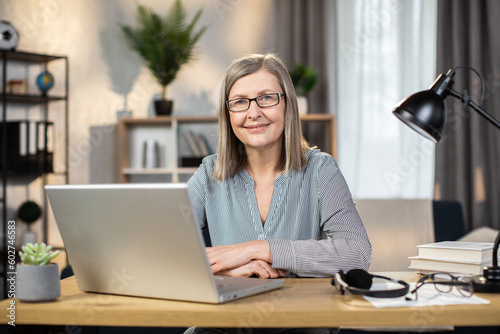 Obraz na plátně Portrait of happy mature lady in everyday clothes posing with laptop on writing desk in cozy workplace indoors