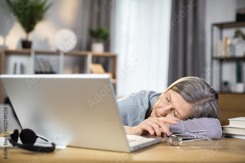 Elderly caucasian woman falling asleep at writing desk with portable computer and glasses aside. Tired female entrepreneur getting overworked and stressed out while doing full-time job at home.